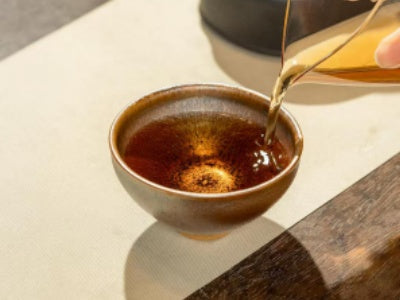 Pouring amber tea into a handcrafted Jian Zhan cup - golden crystalline oil spot glaze catching the light, Jingdezhen tenmoku ware