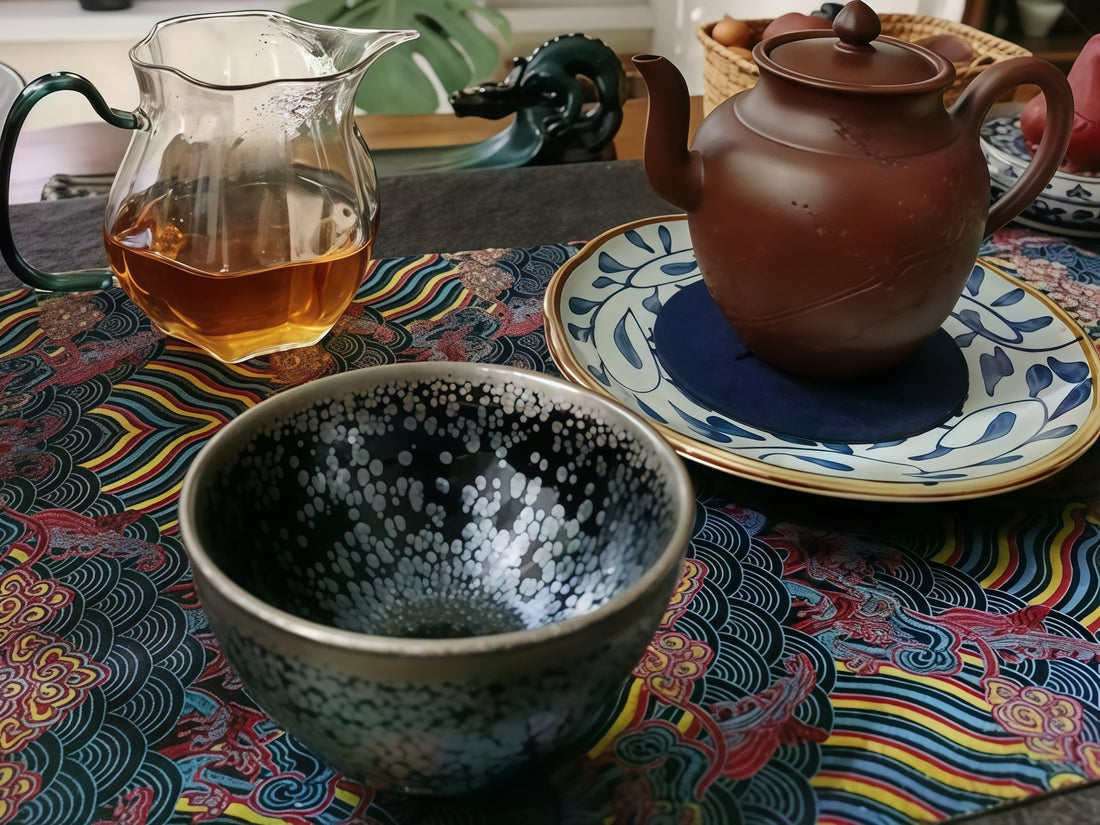 Jian Zhan tenmoku bowl with silver oil spot glaze on an embroidered tea cloth, yixing teapot and glass pitcher behind